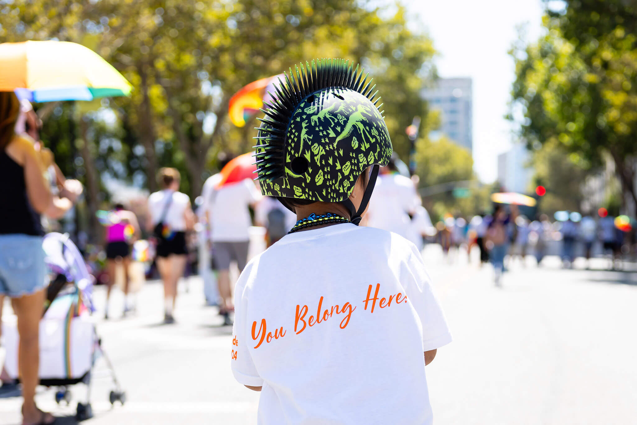 Child wearing a helmet participating in LGBTQIA+ parade wearing a shirt that says "You Belong Here"