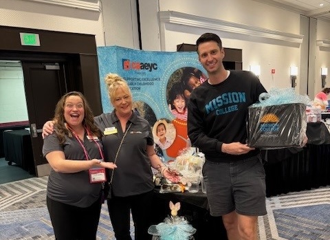 Three people smiling at a conference booth; one holds a gift basket and wears a “Mission College” sweatshirt.