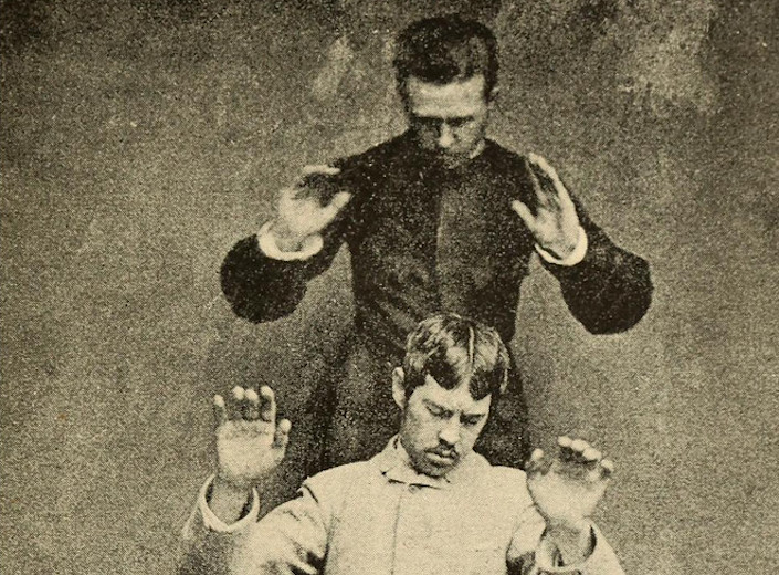 Telepathy. An old photo of a man with his hands up in the air. He is standing behind another man who looks down with his eyes closed in concentration.